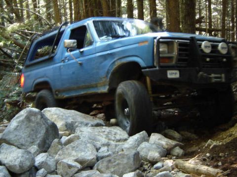 1985 Ford Bronco - A blue Ford Bronco is navigating a rocky, wooded trail. The off-road vehicle is angled towards the viewer's left, with its front tire clear…