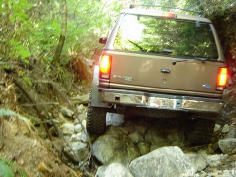 1985 Ford Bronco - The rear of a tan Ford Explorer is navigating a rocky, wooded trail. The vehicle's brake lights are illuminated as it drives over large roc…