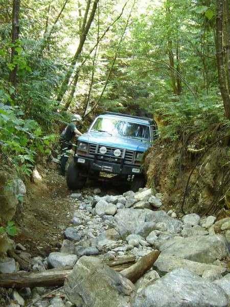 1985 Ford Bronco - A blue 1985 Ford Bronco is navigating a rocky, wooded trail. The vehicle is angled towards the right, with its front grille and headlights…