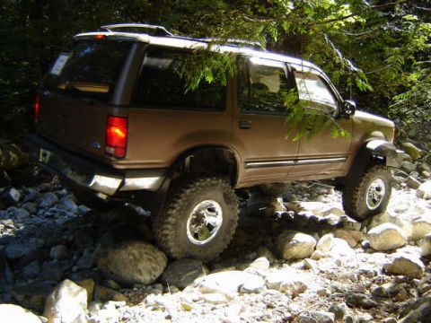 1985 Ford Bronco - A brown Ford Explorer SUV is navigating a rocky, wooded trail, with its tires positioned on large stones.