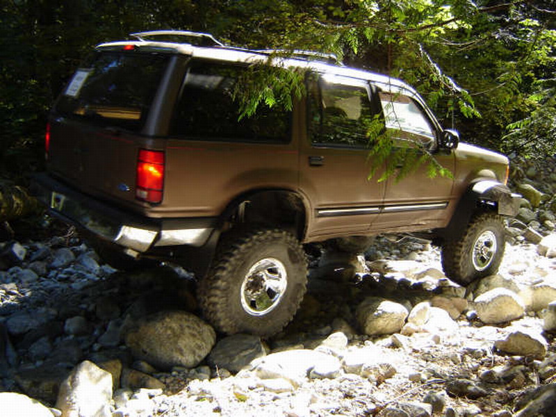 Brown Ford Explorer SUV off-roading on a rocky trail surrounded by trees.