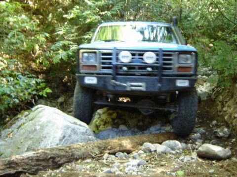 1985 Ford Bronco - A blue Ford Bronco is navigating a rocky, wooded trail, with its front tires positioned over large rocks and logs. The vehicle features a b…