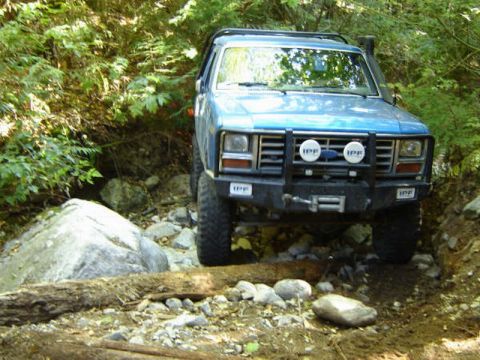 1985 Ford Bronco - A blue 1980s Ford Bronco is navigating a rocky, wooded trail. The off-road vehicle features a prominent aftermarket bumper with integrated…