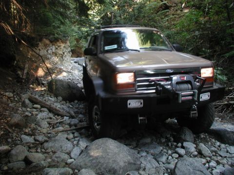 Tan SUV with winch driving over rocks on a wooded trail.