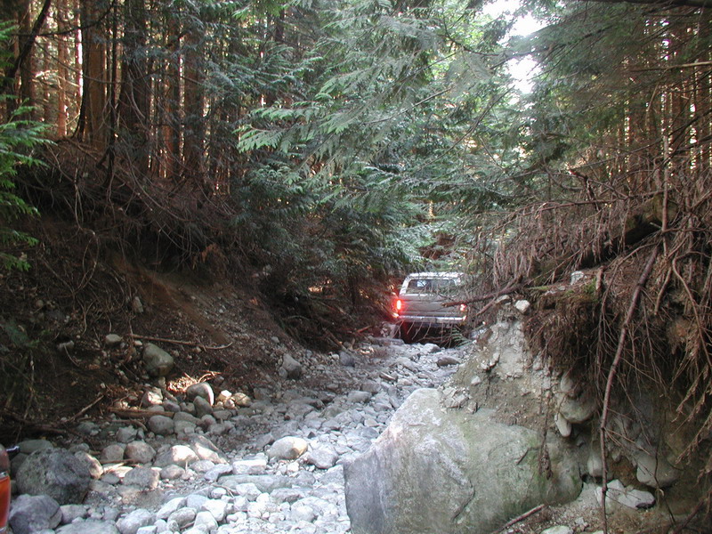 White pickup truck driving on a rocky, wooded trail.