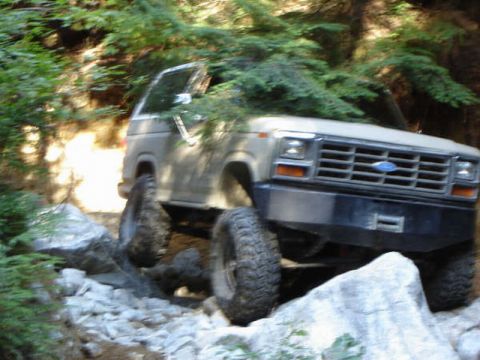 Tan 1980s Ford Bronco off-roading on a rocky, wooded trail.