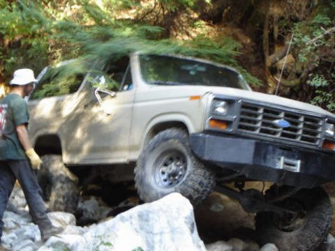 Tan 1980s Ford Bronco off-roading on a rocky trail with a person standing nearby.