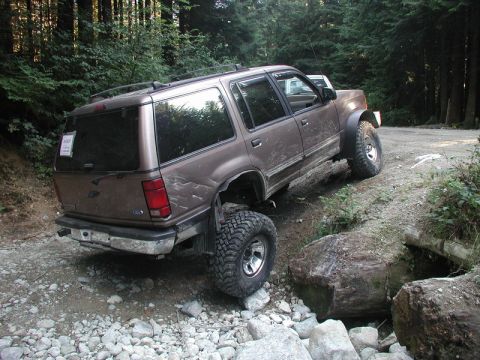 Brown Ford Explorer off-roading on a rocky, wooded trail, with one tire elevated.