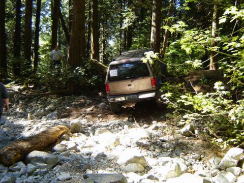 Tan Ford Bronco parked on a rocky, wooded trail with a person in the background.