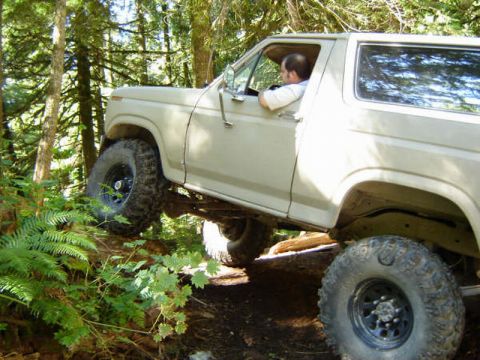 Tan 1980s Ford Bronco driving over a log on a wooded trail.
