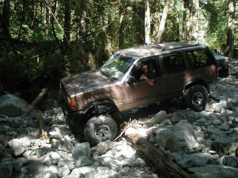 Lifted brown SUV driving over rocks in a forest.