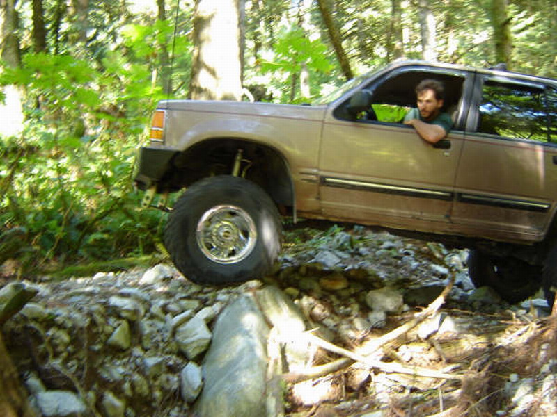 Tan SUV off-roading on a rocky trail in a forest, driver visible.