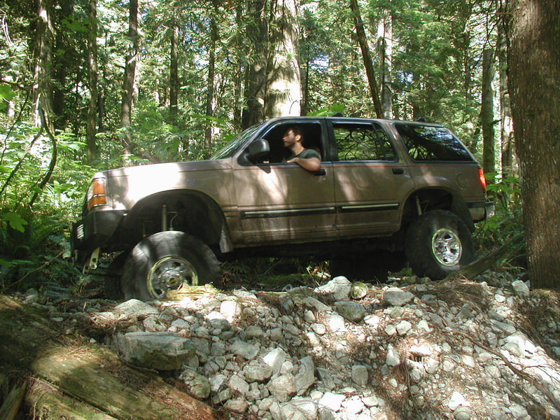 Lifted tan Ford Explorer off-roading on a rocky trail in a forest.