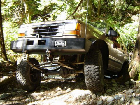 Brown SUV with aftermarket bumper and off-road tires on a rocky trail in a forest.