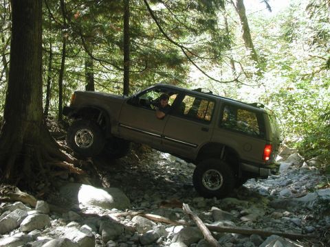 Lifted tan Ford Explorer off-roading on a rocky, wooded trail.
