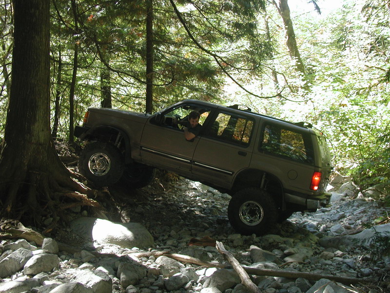Lifted tan Ford Explorer off-roading on a rocky, wooded trail.