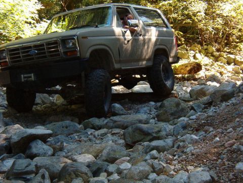 Tan 1980s Ford Bronco driving over rocks on a wooded trail