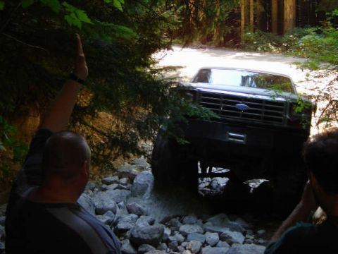 Dark Ford Bronco off-roading on a rocky trail surrounded by trees, person's arm raised.