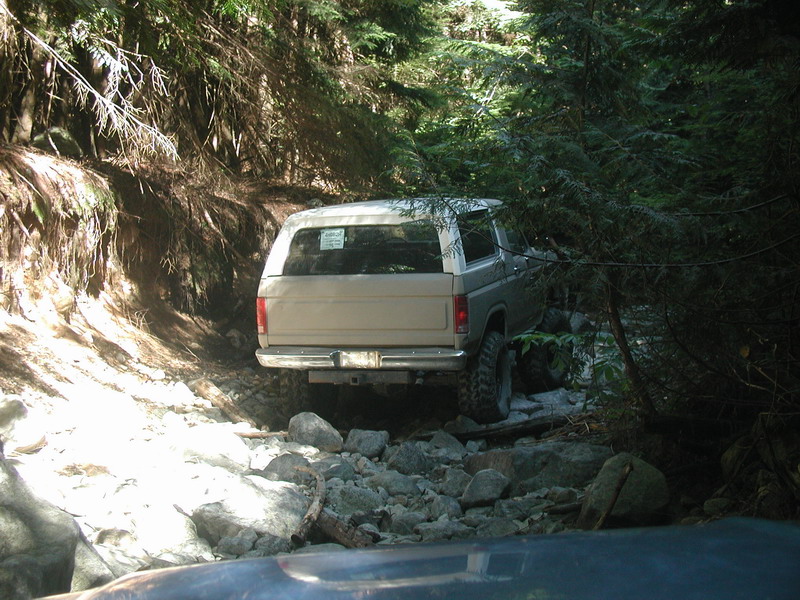 Tan Ford Bronco driving on a rocky, wooded trail.