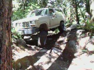 Tan 1980s Ford Bronco off-roading on a rocky trail, front passenger tire lifted.