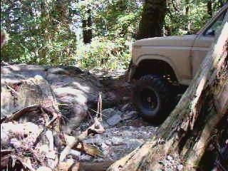 Tan 1985 Ford Bronco on a rocky, wooded trail, front passenger side view.