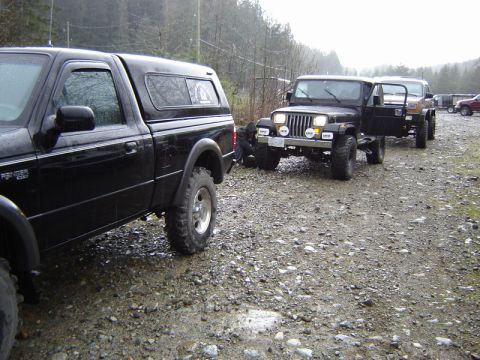 1985 Ford Bronco - A line of off-road vehicles, including a black Ford Ranger pickup truck and several Jeep Wranglers, are parked on a gravel road in a wooded…