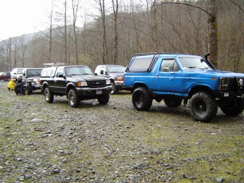 1985 Ford Bronco - A blue Ford Bronco with off-road modifications is parked on a gravel lot in a wooded area. Several other trucks and SUVs are parked behind…