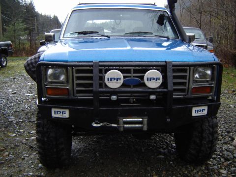 1985 Ford Bronco - Front view of a blue Ford Bronco with a black bull bar, winch, and IPF lights. The vehicle is parked on a gravel surface with trees in the…