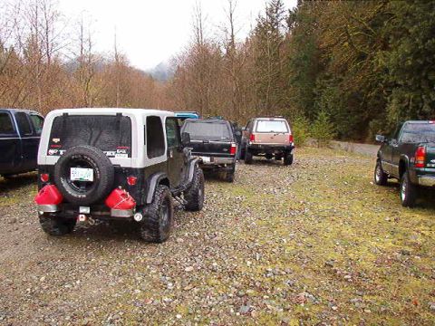 1985 Ford Bronco - The rear of a two-tone black and silver Jeep Wrangler is shown parked on a gravel lot with trees in the background. Two red jerry cans are…