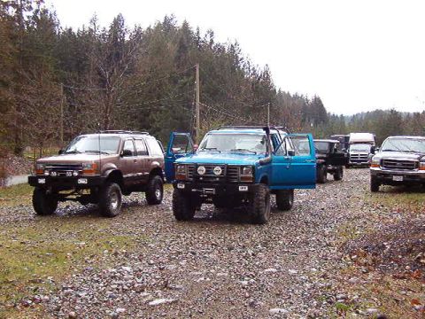 1985 Ford Bronco - A lineup of modified SUVs and trucks are parked on a gravel road with a wooded background. The blue SUV in the foreground has its driver's…