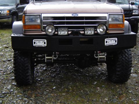1985 Ford Bronco - The front view of a modified Ford Bronco shows a black custom bumper with two Hella decals and multiple tow hooks. The vehicle has large of…