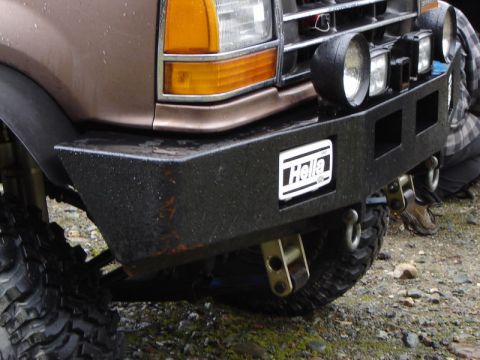 1985 Ford Bronco - Close-up of the front bumper and fender of a brown vehicle, featuring a black aftermarket bumper with a Hella logo and auxiliary lights mou…