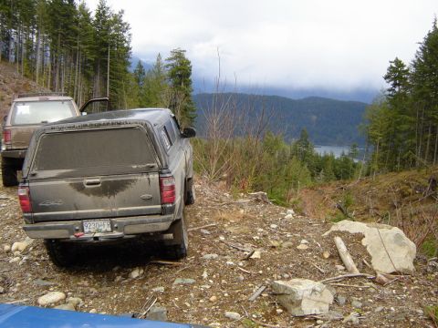 1985 Ford Bronco - Two pickup trucks are parked on a dirt road overlooking a lake and forested mountains. The foreground truck is a dark-colored Ford Ranger w…