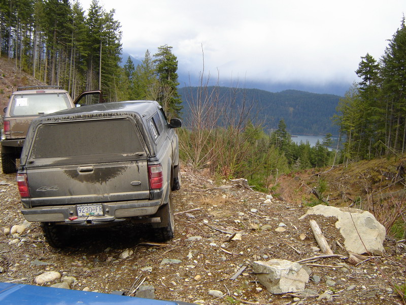 Two pickup trucks on a dirt road with a lake and mountains in the background.