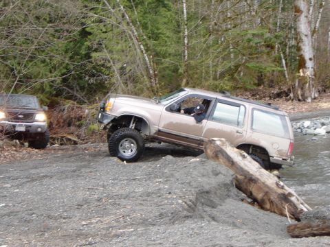 1985 Ford Bronco - A lifted tan SUV is navigating a rocky stream bed with a large log in the foreground. Another dark-colored SUV is visible in the background.