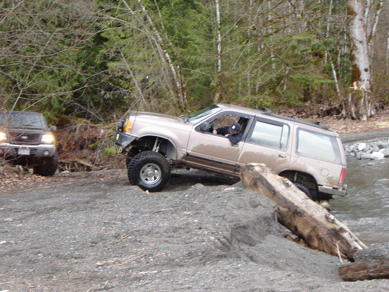 Lifted tan SUV driving through a rocky stream with a log in the foreground.