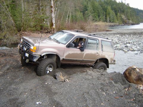 1985 Ford Bronco - A tan Ford Explorer is fording a shallow stream on a rocky shore, with its front tires partially submerged in sand and mud.