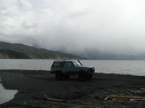 1985 Ford Bronco - A lifted, dark teal Ford Bronco is parked on a dark, pebbly beach next to a large body of water. The sky is overcast with heavy clouds obsc…