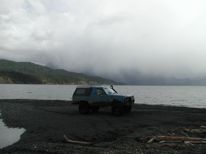 Lifted dark teal Ford Bronco on a beach with overcast skies and mountains in the background.