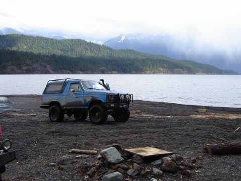 1985 Ford Bronco - A lifted, blue 1985 Ford Bronco is parked on a dark, rocky beach next to a large body of water. The vehicle has a snorkel, a brush guard, a…