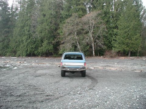 1985 Ford Bronco - The rear view of a blue Ford Bronco parked on a gravel beach with a dense forest in the background.