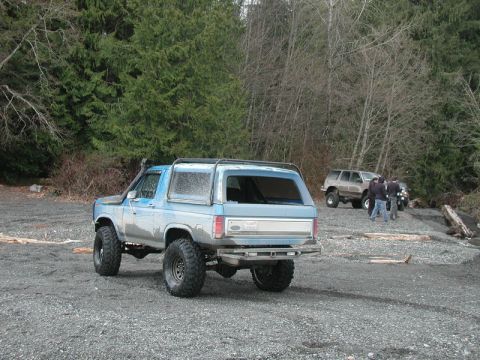 1985 Ford Bronco - A lifted blue Ford Bronco with large off-road tires and a snorkel sits on a gravel beach next to a forest. Other SUVs and people are visibl…