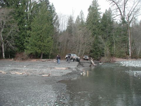 1985 Ford Bronco - Two SUVs are parked on a gravelly shoreline next to a river, with a dense forest in the background. One SUV is a dark-colored Jeep Wrangler…