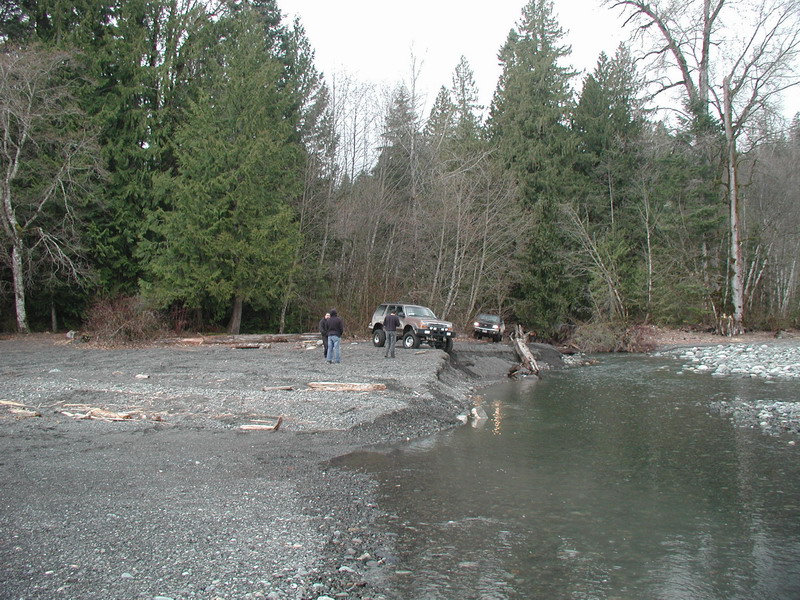 Two SUVs parked on a riverbank with a forest behind them.