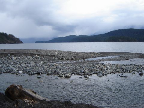 1985 Ford Bronco - A wide shot of a rocky shoreline with a body of water and forested hills in the background under a cloudy sky.