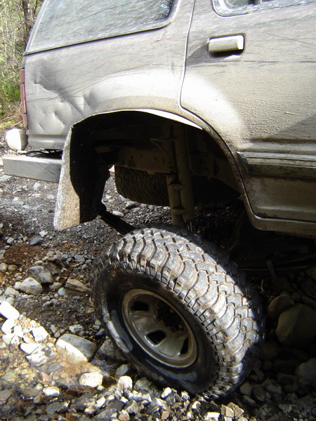 1985 Ford Bronco - The rear quarter panel and wheel of a muddy, dented SUV are shown off-road. The vehicle is positioned on a rocky terrain with some water fl…