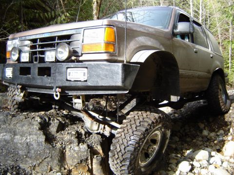 1985 Ford Bronco - A lifted brown Ford Explorer is navigating a rocky, off-road trail with its front tire on a large rock. The vehicle features a custom black…