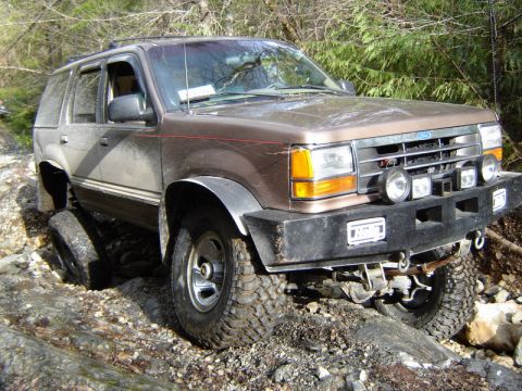 1985 Ford Bronco - A brown Ford Explorer is navigating a rocky, off-road trail with its front passenger side tire elevated.
