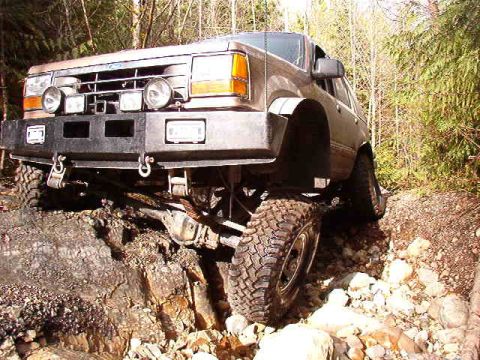 1985 Ford Bronco - A modified brown SUV is navigating a rocky, wooded trail, with its front tire angled up a large rock.