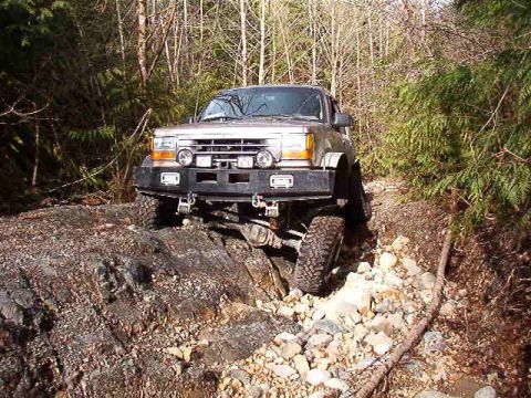 1985 Ford Bronco - A silver Ford Bronco is navigating a rocky, wooded trail. The vehicle is angled towards the right, with its front left tire on a large rock.